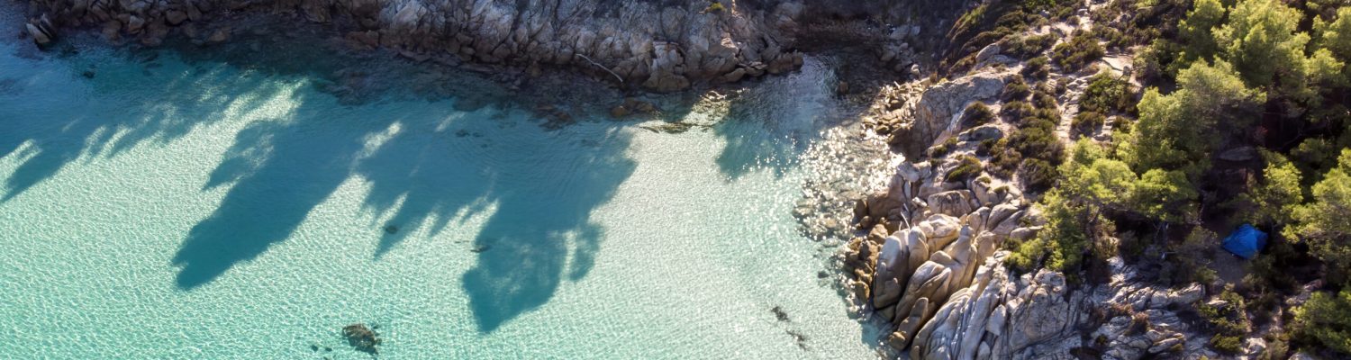 Aegean sea coast with blue transparent water, greenery around, rocks, bushes and trees, view from the drone, Greece